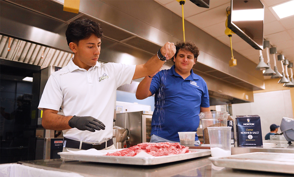 Man in white shirt sprinkles seasoning on meat in a kitchen classroom.