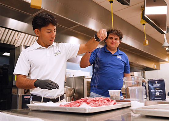 Man in white shirt sprinkles seasoning on meat in a kitchen classroom.