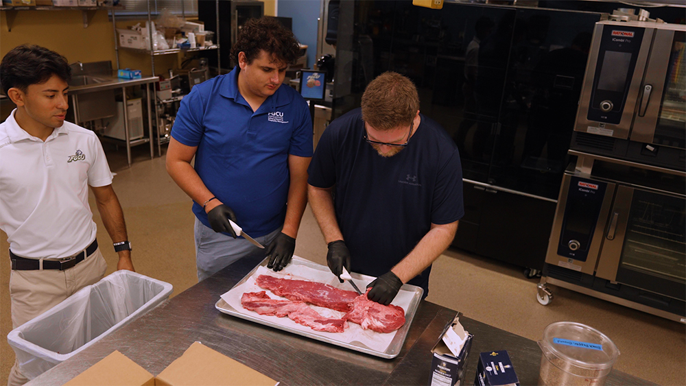 Three men in kitchen classroom trimming meat with knives.