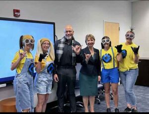 Six people, four in minion costumes, one man in a Gru costume, and one blonde woman in a skirt and black blazer.