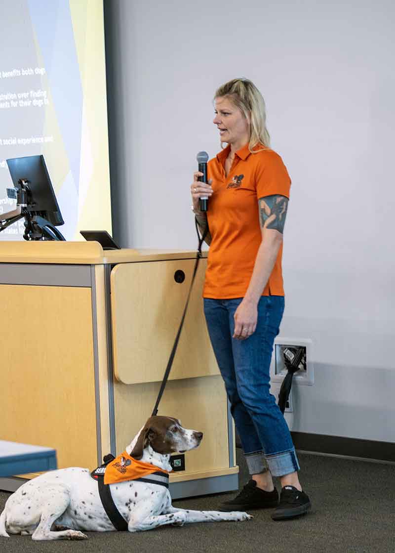 A woman in an orange polo and jeans talks into a microphone, while her leashed white and brown dog sits at her feet