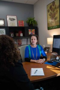 A woman in a blue and green shirt sits behind a desk, talking to a young woman