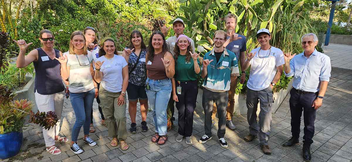 A panoramic photo of one dozen people outdoors, holding up the FGCU Wings Up gesture