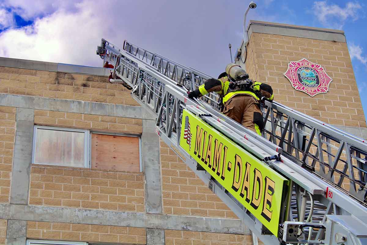 A fire fighter climbs an aluminum fire ladder to the roof of a building. The words Miami Dade are on the ladder