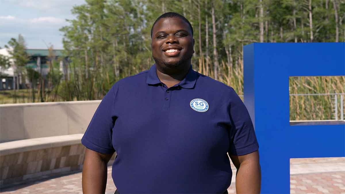 A man in a dark blue polo with the FGCU Student Government logo stands outdoors near the FGCU logo on the Library Lawn