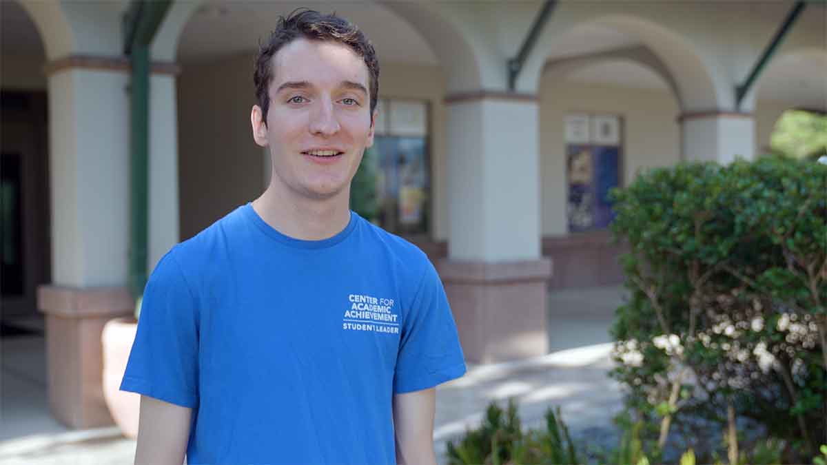A man in a blue t-shirt with an FGCU Center for Academic Achievement Student Leader logo stands outdoors