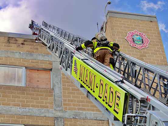 A fire fighter climbs an aluminum fire ladder to the roof of a building. The words Miami Dade are on the ladder