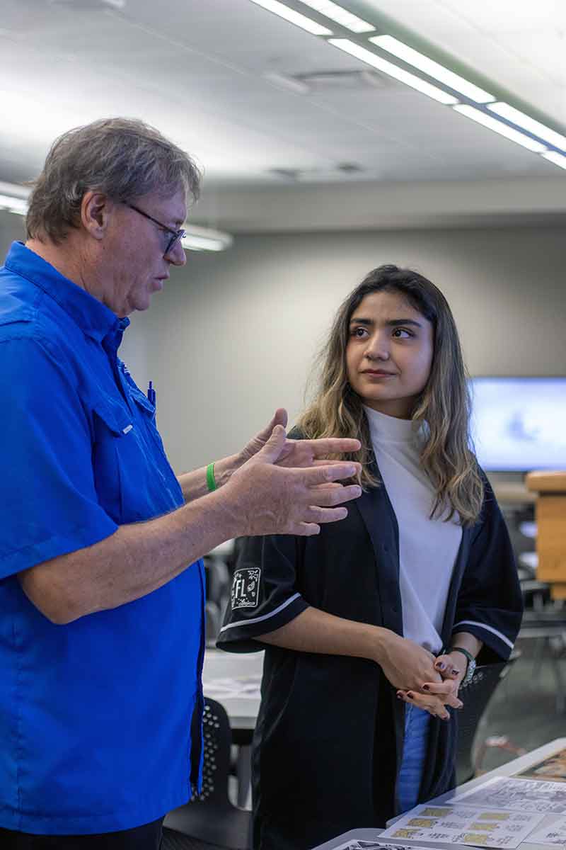 A man in a bright blue button-down shirt talks to a young woman in a black shirt worn over a white shirt
