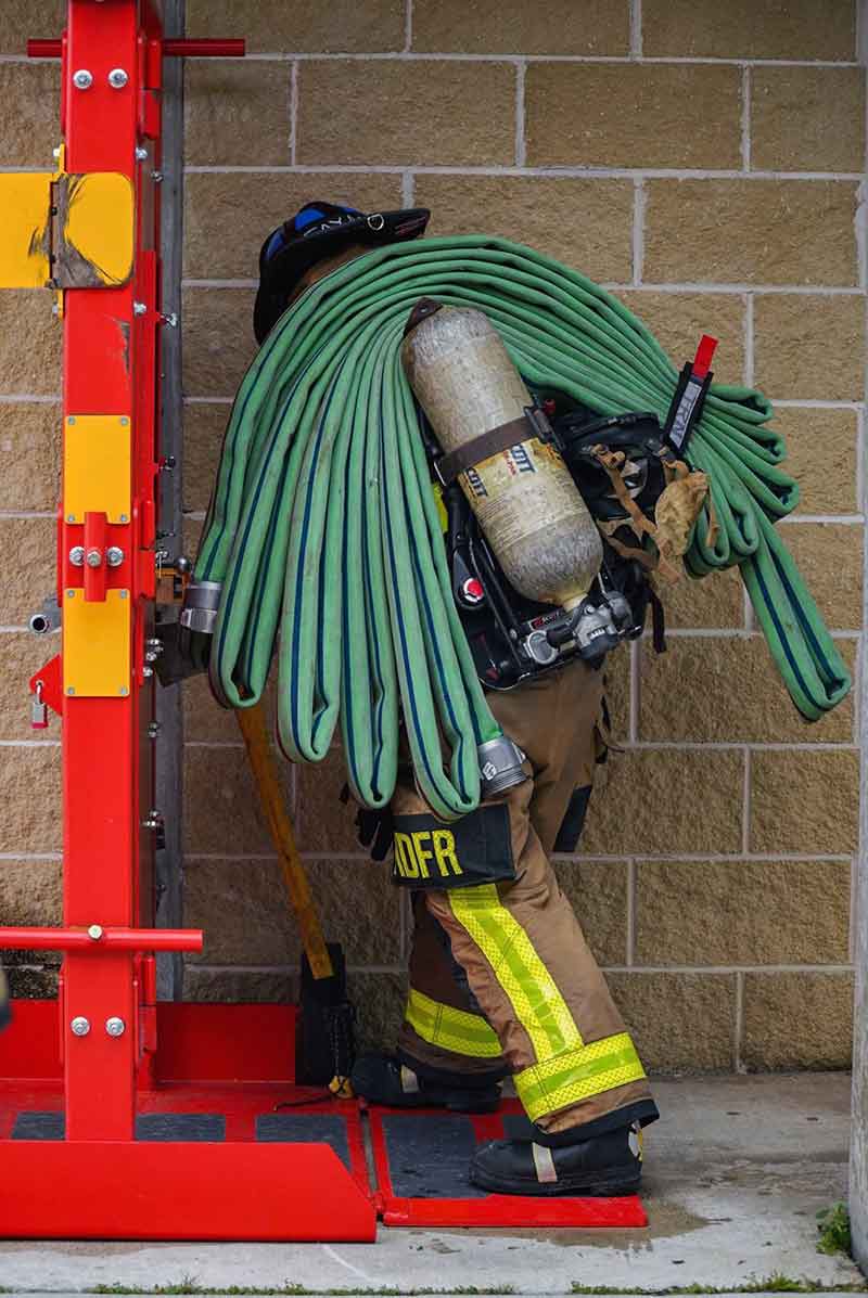 A firefighter in full gear stands before a vertical ladder with a green firehose on her shoulder