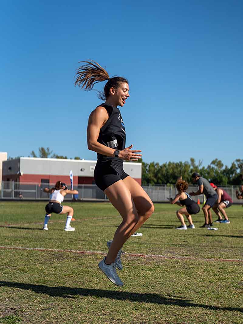A woman with toned arms and legs in a black sleeveless top and bike shorts is mid-jump, with her ponytail flying in the air