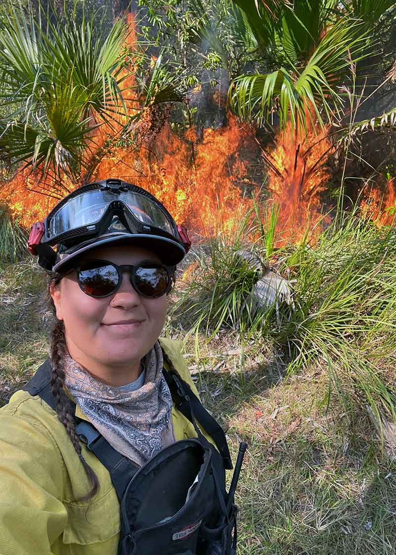 A woman in protective gear and clothing standing in front of a bush on fire during a prescribed burn