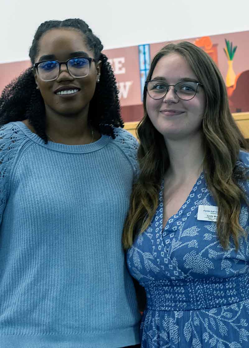 A Black woman in a blue sweater and a white woman in a blue dress, both wearing glasses and smiling, in front of a display of books