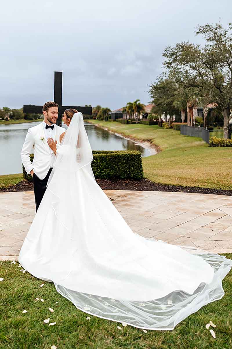 A man in a white tuxedo jacket, black pants, black bow-tie and white shirt stands with his arm around a bride in a white dress, mesh veil and long train, outdoors and in front of a small pond and large Christian cross