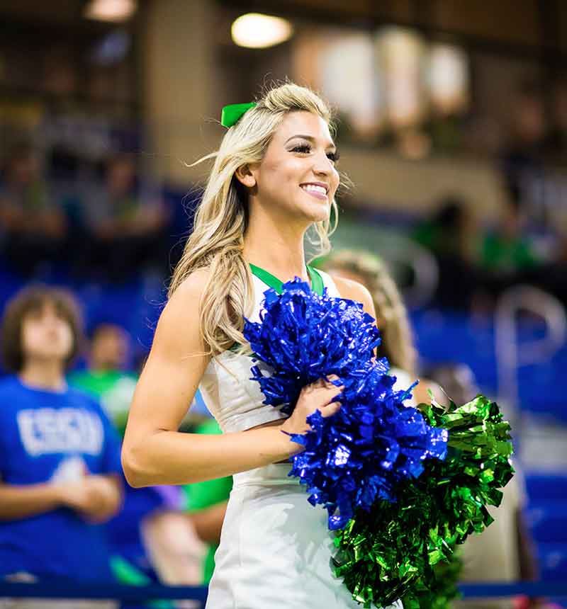 A young blonde woman in a white cheerleader top and skirt trimmed in green holds two pom-poms, one FGCU blue and one FGCU green