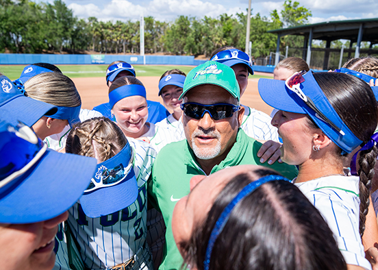 FGCU softball coach in green shirt and hat surrounded by his players wearing blue