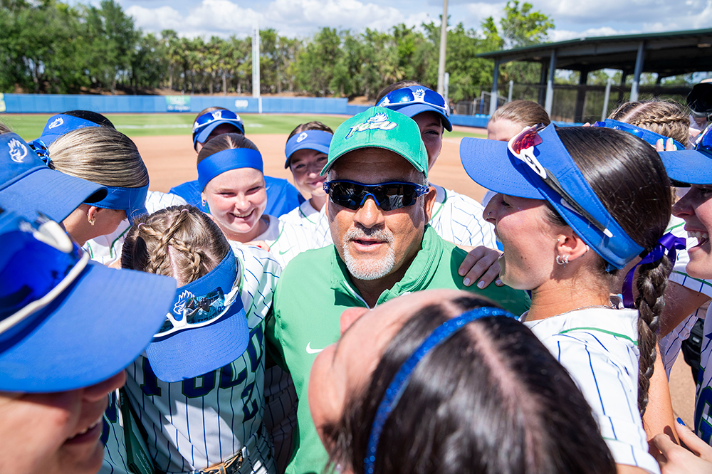 FGCU softball coach in green shirt and hat surrounded by his players wearing blue