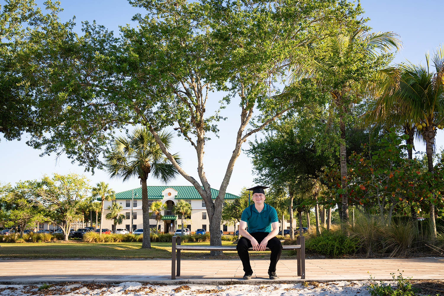 Man in graduation cap, teal shirt and black pants sitting on a bench under a tree