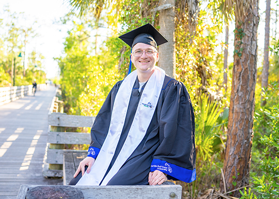 Man in graduation cap and gown sitting on boardwalk bench