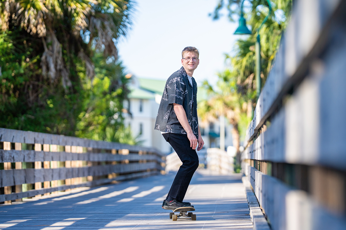 Man with glasses wearing a. black shirt and riding a skateboard on a boardwalk.