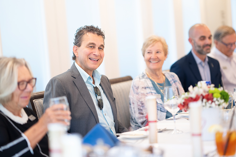 man in gray suit with shirt sits at a table with guests at a luncheon