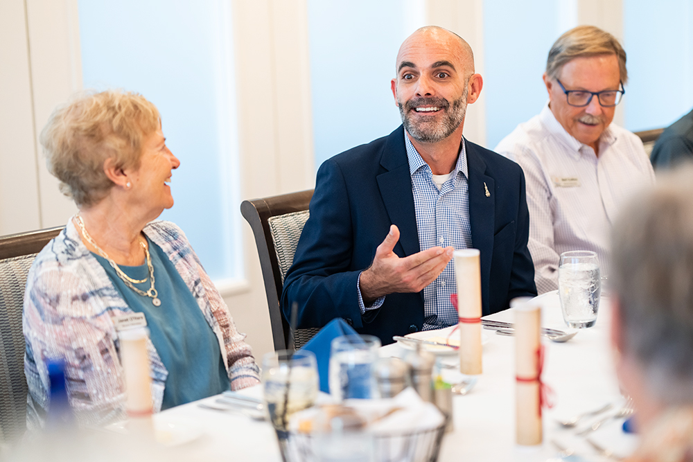man in dark blue suit with sits at a table with guests at a luncheon