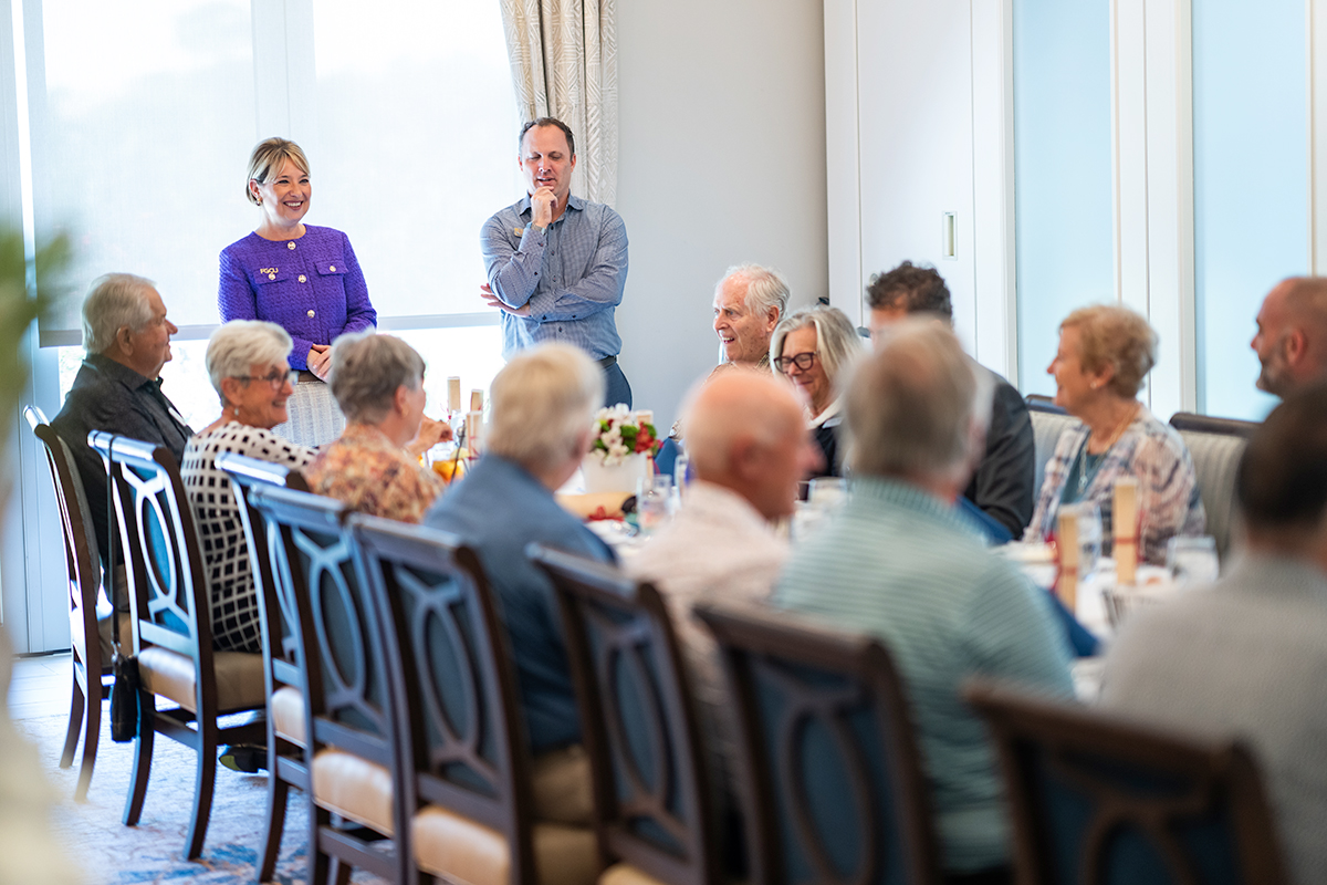 A woman in a purple suit and a man in a blue shirt stand at the head of table where people sit