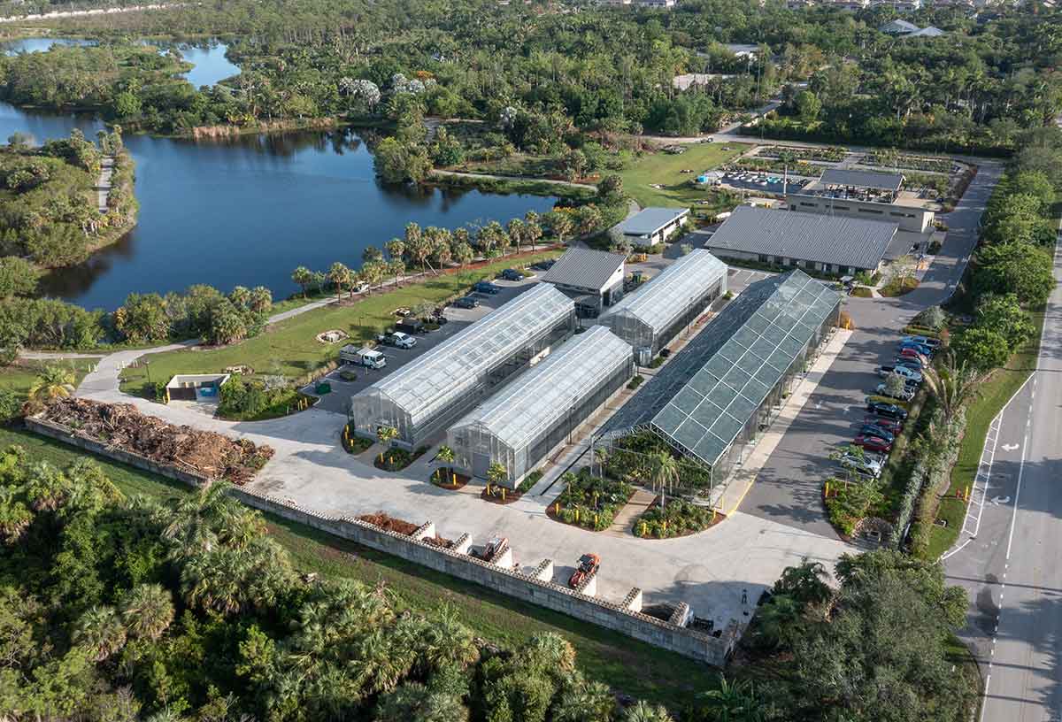An aerial view of large greenhouses and buildings in rows with a large pond on the left, a paved road on the right and forested areas surrounding