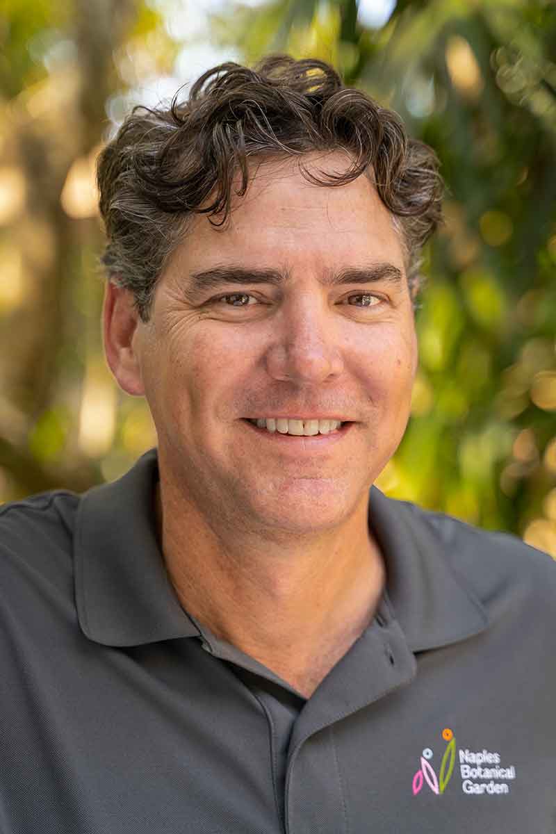 A smiling man in a grey polo shirt with a blurry background of green nature