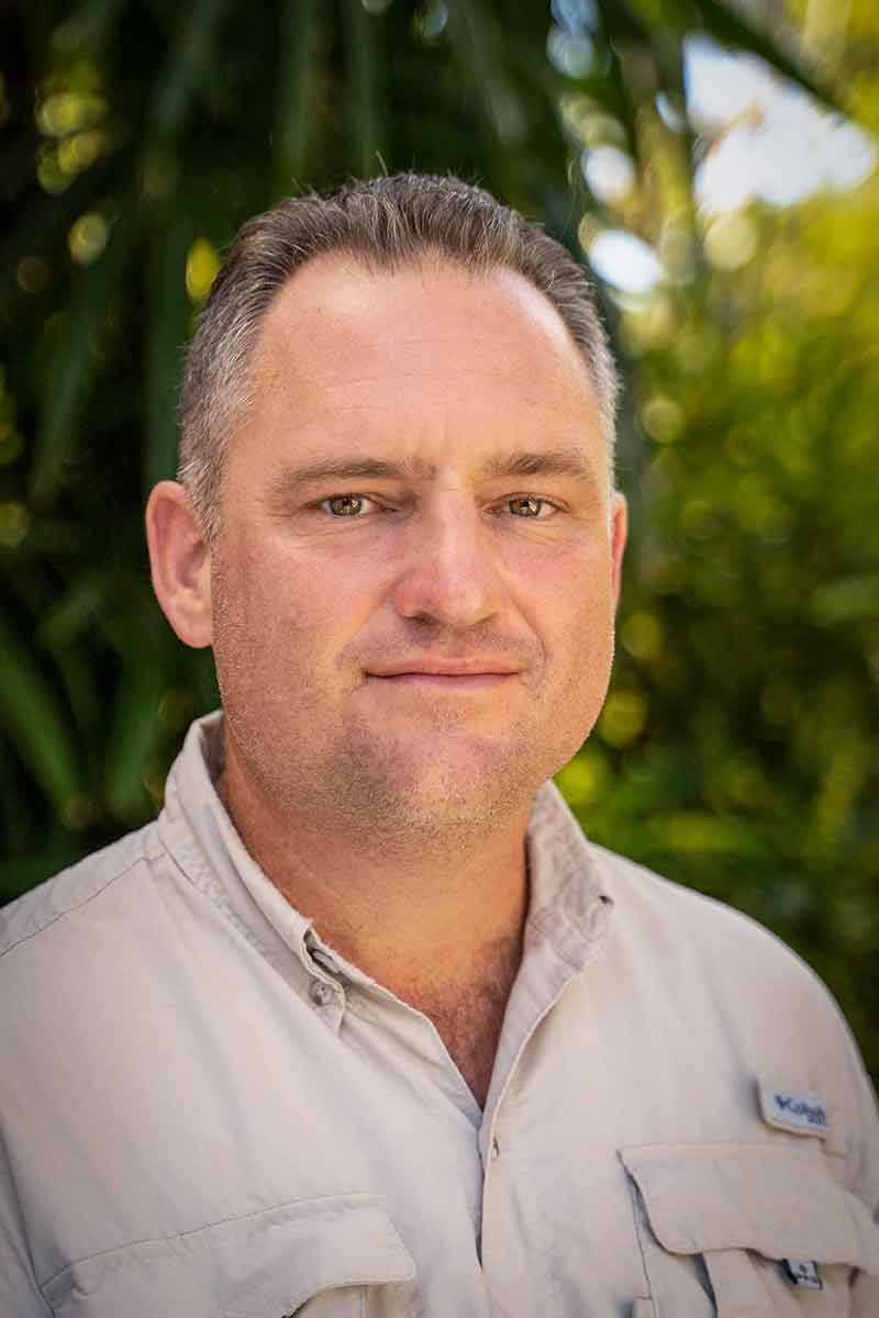 A man in a beige work shirt poses in front of a green natural landscape