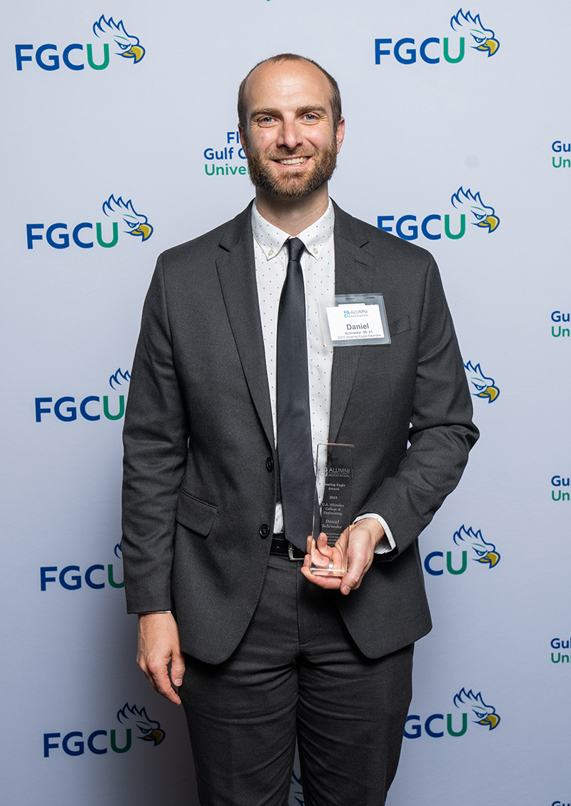 A person wearing a dark suit, white shirt with small black dots, and a black tie stands in front of a backdrop with the FGCU logo