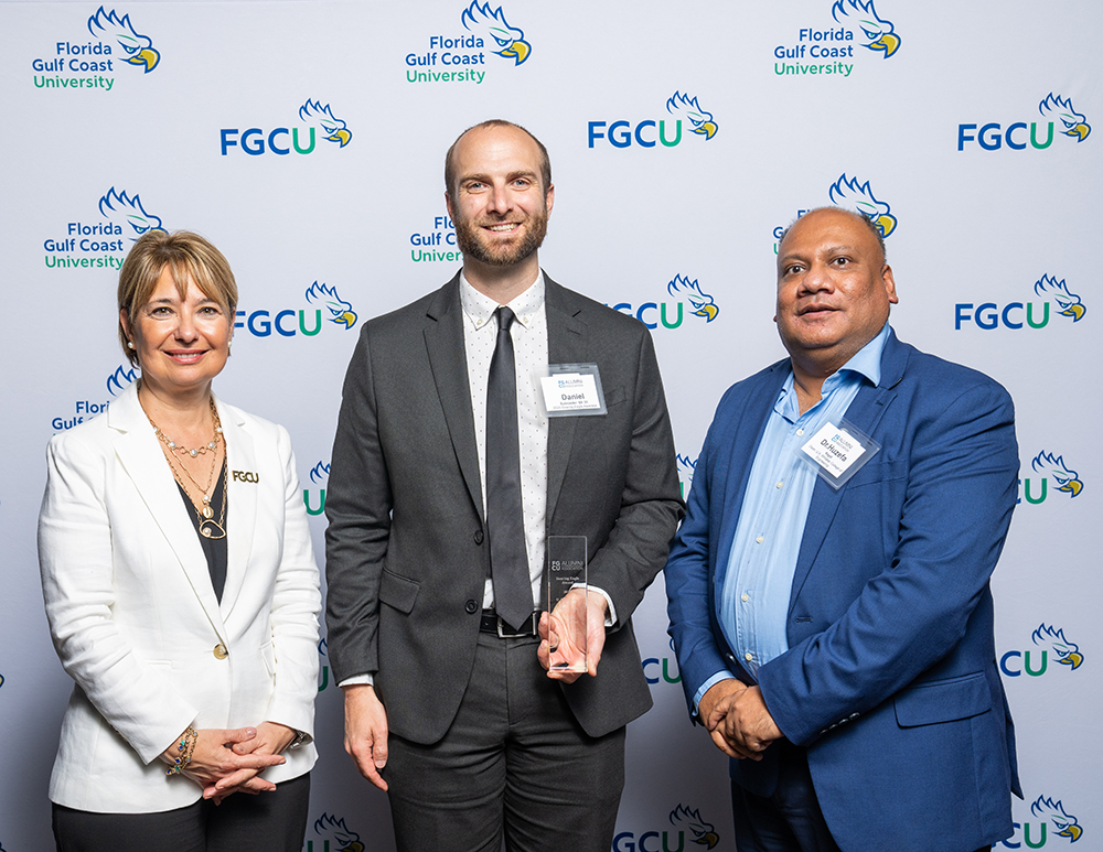 Three individuals stand in front of a Florida Gulf Coast University backdrop. The center person holds an award.