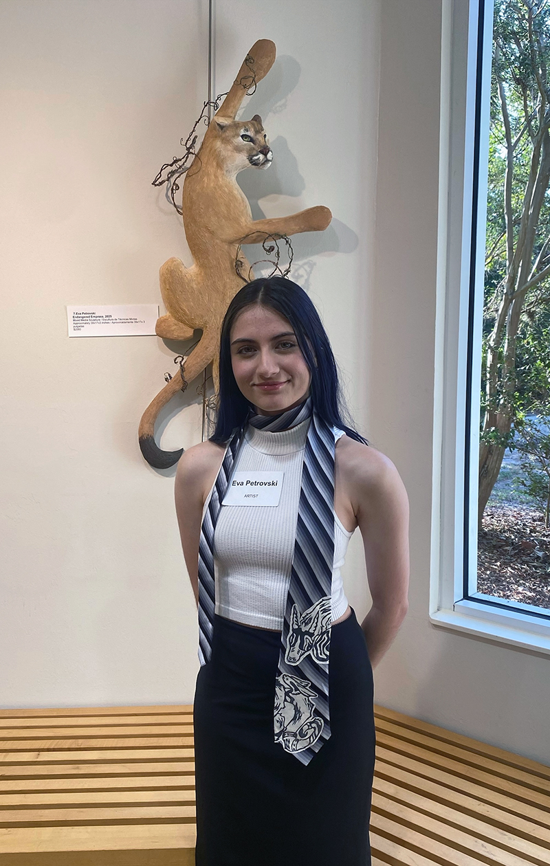 A person with long dark hair stands in front of a wall-mounted puma sculpture titled "The Prowess." They wear a white sleeveless top, black skirt, and an animal-themed striped tie.