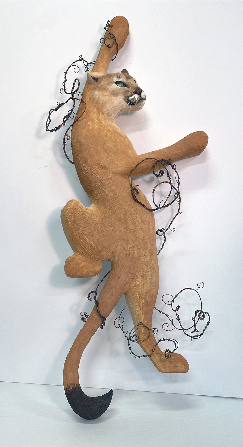 A sculpted mountain lion is mounted on a white wall, shown mid-leap with front paws extended upward and back legs stretched behind. The muscular form and facial features are detailed. Thin black wires wrap around its body and limbs, adding an abstract element. The tail curves downward with a black tip.