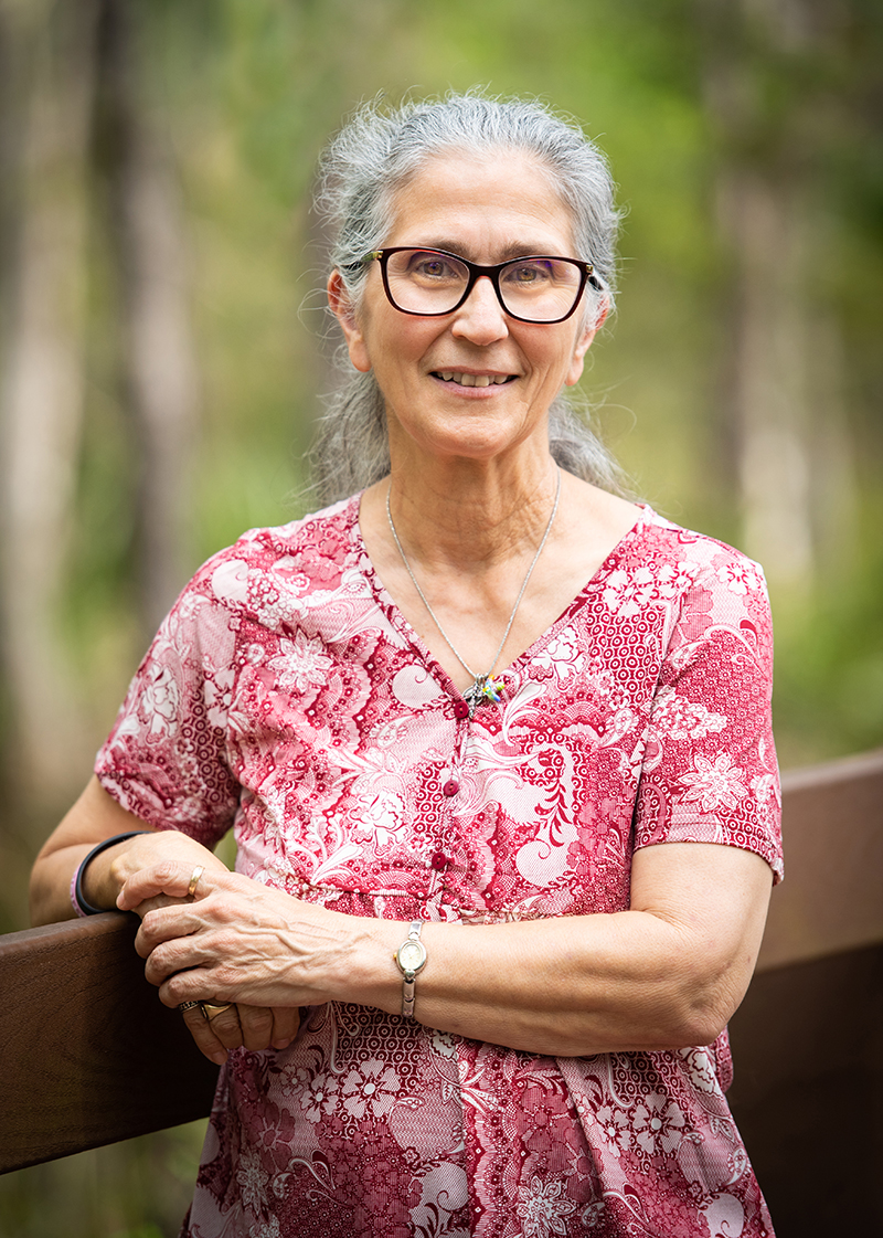 An older woman with silver-white hair and dark-rimmed glasses in a pink v-neck shirt stands on a boardwalk with a blurry image of green trees behind her
