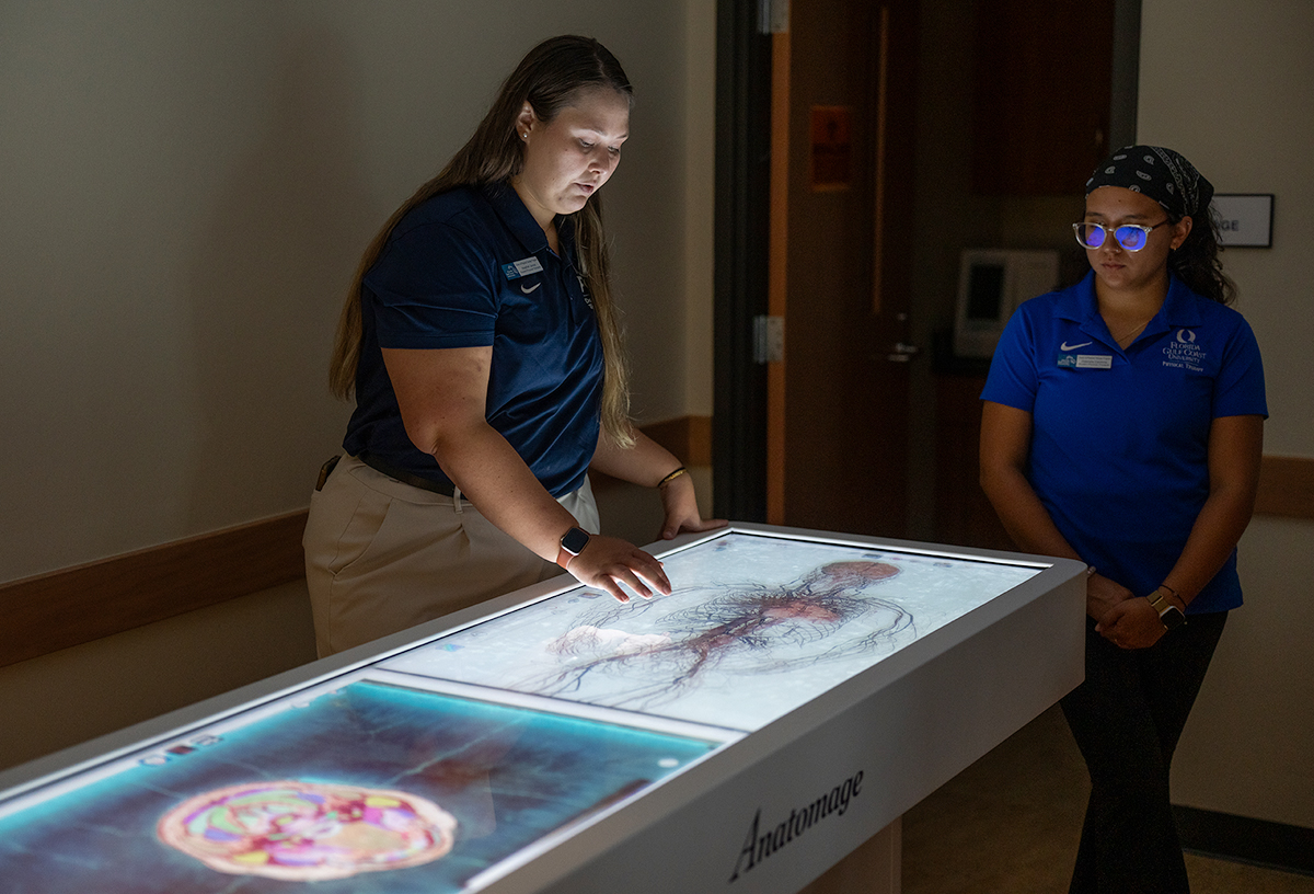 Two individuals in uniforms stand in a room, with one interacting with an Anatomage table displaying a cross-sectional human brain and circulatory system. Both wear name tags, indicating an educational setting.