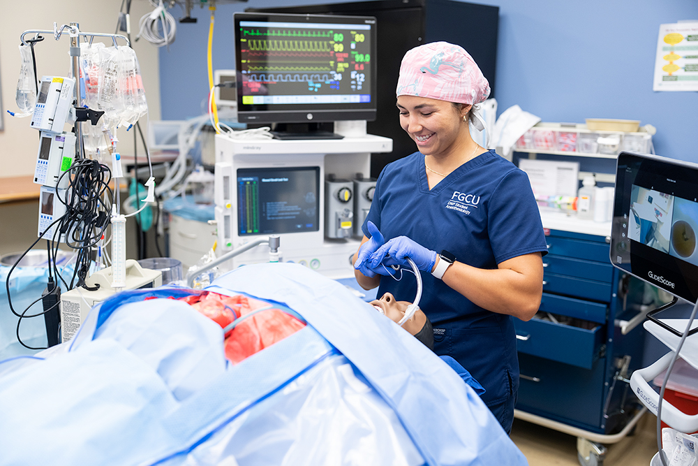 A student in blue scrubs and a pink surgical cap performs a procedure on a simulated patient covered with blue surgical drapes. Surrounding them are medical devices including an anesthesia machine, IV stands, and monitors displaying vital signs.