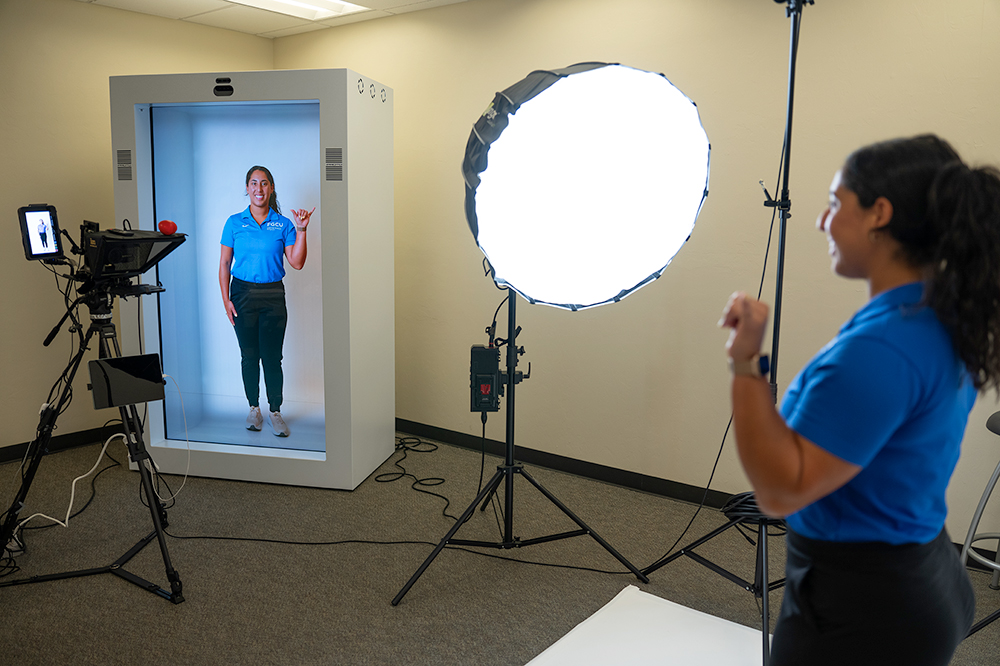 A person in a blue shirt and black pants poses inside a white booth with a transparent front, making a peace sign. In front of the booth is professional photography equipment, including a ring light, camera on tripod, and iPad.