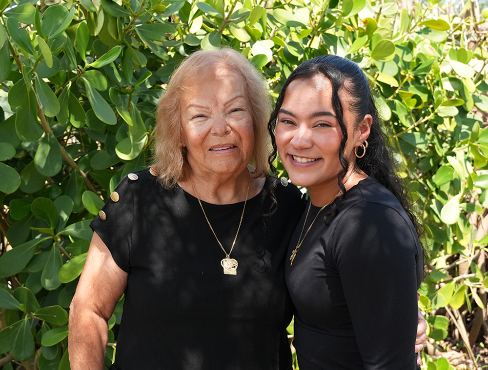 An older blonde woman in a black t-shirt and a younger woman with long black hair and a long-sleeve black shirt stand smiling outdoors