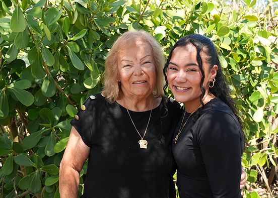 An older blonde woman in a black t-shirt and a younger woman with long black hair and a long-sleeve black shirt stand smiling outdoors