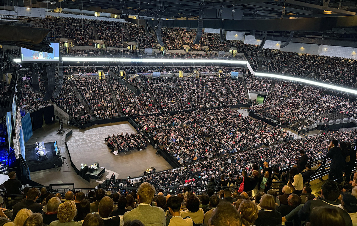 A large indoor arena packed with a massive audience. The stage is on the left, with two large screens displaying the event. Multiple tiers of seating and bright lighting fill the space.