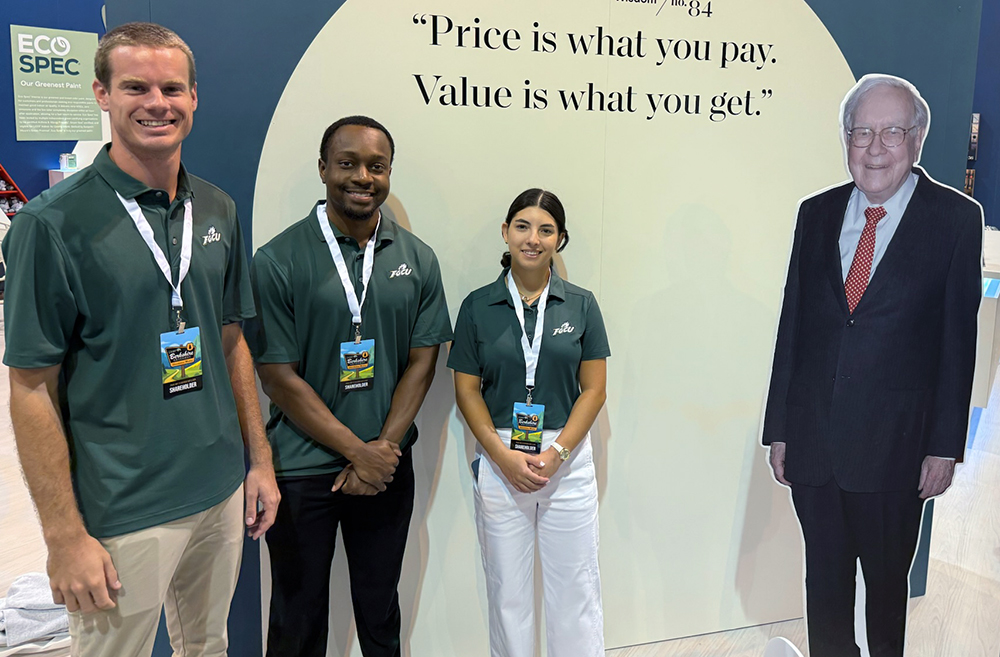 Three people in green polo shirts with name badges stand beside a life-sized cardboard cutout of Warren Buffett. Behind them is a large sign displaying the quote, 'Price is what you pay. Value is what you get.' An 'ECO SPEC' sign is visible on the left.