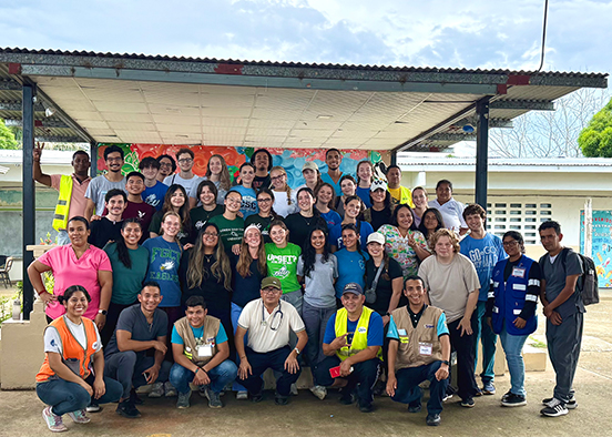 A large group of approximately 40 people pose for a group photo under a covered outdoor area. They are arranged in several rows, some standing and others kneeling. The group includes individuals in casual clothing, uniforms, and safety vests, with some wearing badges or lanyards. Behind them is a colorful mural on the wall, part of a building with windows, and another mural visible to the right.
