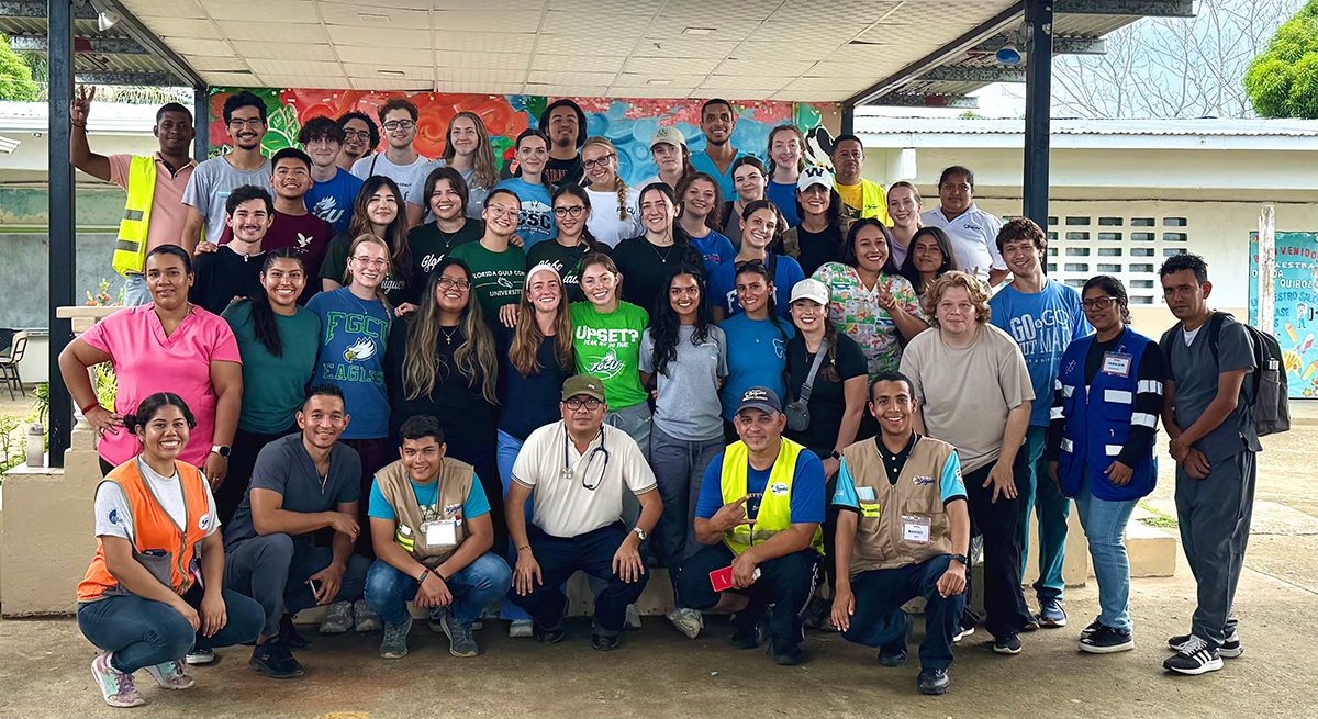 A large group of approximately 40 people pose for a group photo under a covered outdoor area. They are arranged in several rows, some standing and others kneeling. The group includes individuals in casual clothing, uniforms, and safety vests, with some wearing badges or lanyards. Behind them is a colorful mural on the wall, part of a building with windows, and another mural visible to the right.