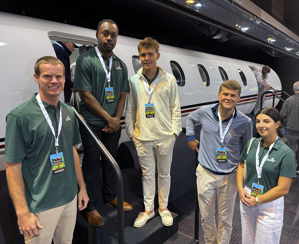 Five people wearing event badges pose in front of a white private jet with red and black stripes at an indoor exhibition. Three wear green polo shirts, two wear light-colored shirts, and one stands on the jet’s staircase