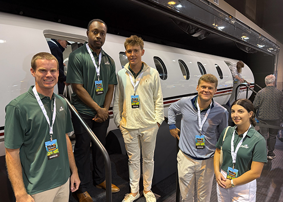 Five people wearing event badges pose in front of a white private jet with red and black stripes at an indoor exhibition. Three wear green polo shirts, two wear light-colored shirts, and one stands on the jet’s staircase