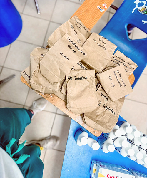 Top-down view of a wooden table with several brown paper bags labeled with medication names and dosages, including 'Ciprofloxacina 500 mg, 20 count,' 'Claritromicina 500 mg, 12 count,' and 'Ceftriaxona 1g, 50 tablets.' White plastic bottles and a box labeled 'CREAT' are on a blue surface next to the table. A person wearing green pants and white shoes is partially visible in the bottom left corner.