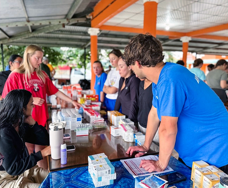 Several people are gathered around a table under a covered outdoor area with orange pillars. The table is filled with medical supplies, including boxes of medication and bottles. One person in a blue shirt is leaning over paperwork on the table, while another in a red shirt is pointing at the supplies. Others are standing or sitting nearby, engaging in conversation or examining the items. Trees and more people are visible in the background.