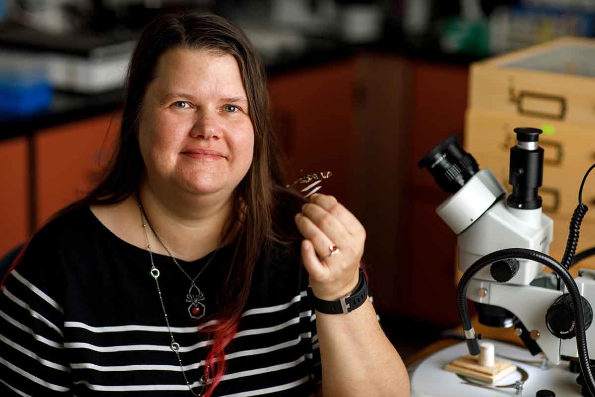 A woman in a black and white-striped shirt holds a small insect and sits in front of a microscope