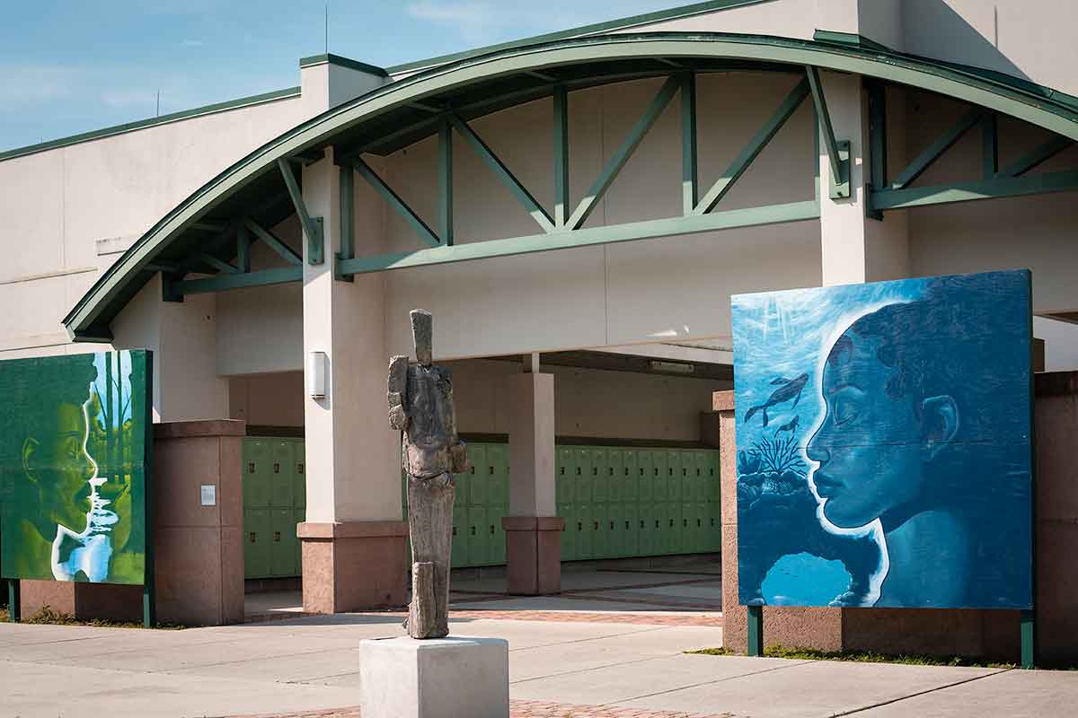 A ceramic statue of a life-sized human figure stands outside a beige academic building with a green roof. Green lockers can be seen inside the corridor of the building, which hosts an oversized green mural of a man's face and an oversized blue mural of a woman's face to either side of the building's entrance