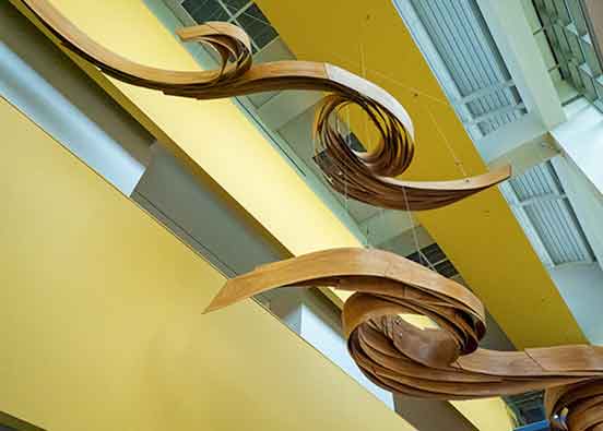 A photo from beneath an art installation hanging from the ceiling. The artwork features two forms, each about 25 feet long, crafted from layered cherry veneer and metal
